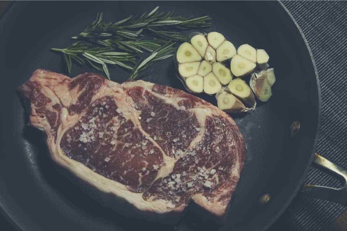 A steak cooking next to rosemary and garlic in a cast iron skillet